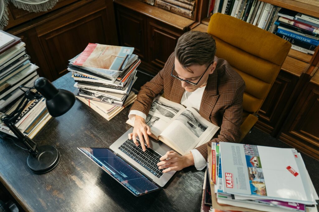 A young man studies in a cozy library setting, using a laptop and books at a wooden desk.
